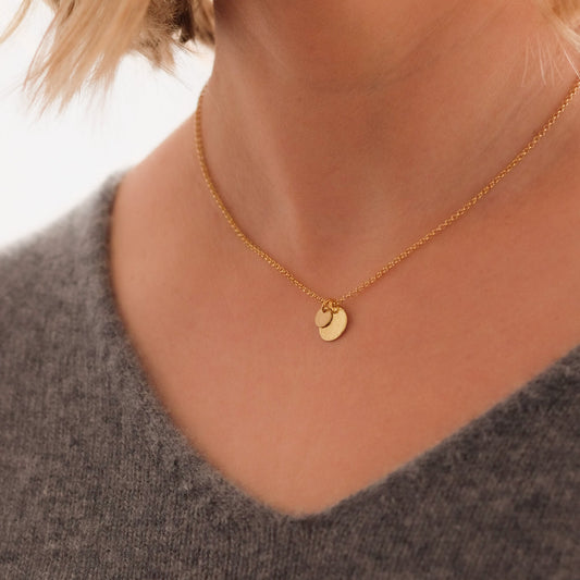 Goldene Kette mit zwei runden Anhängern, um einen Hals liegend. Grauer Pullover mit V-Ausschnitt.